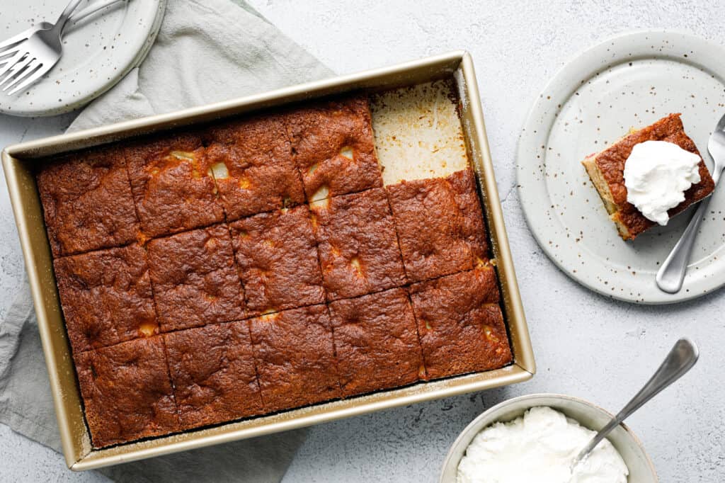 A sheet pan of cake with a slice missing from the pan and placed on a nearby plate, topped with whipped cream.