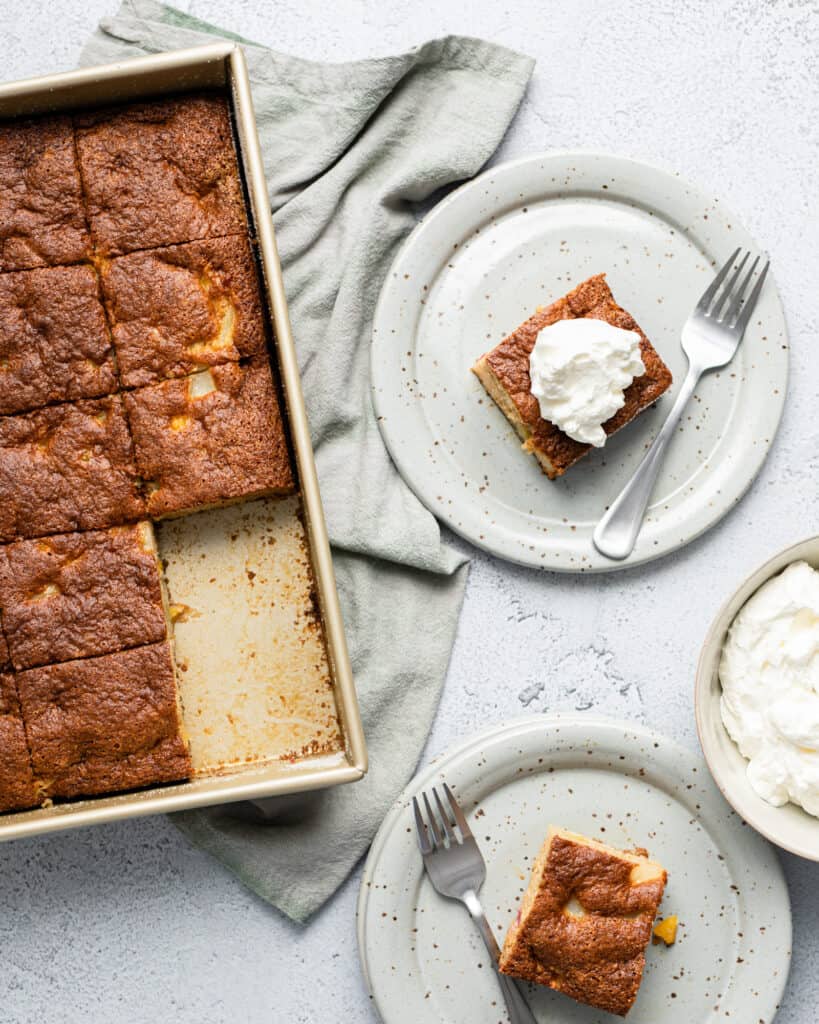 a sheet pan of fruit cocktail cake with two slices cut out and placed onto plates with forks and whipped cream