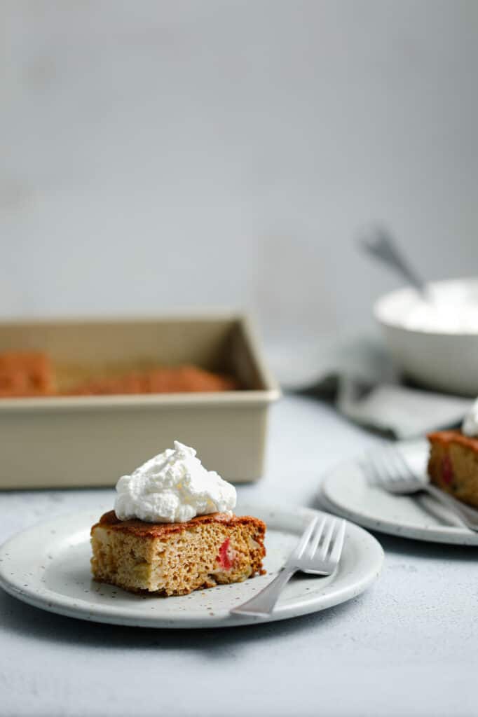 a straight on shot of a slice of square cake with whipped cream on a plate on a grey surface surrounded by plates and the baking dish