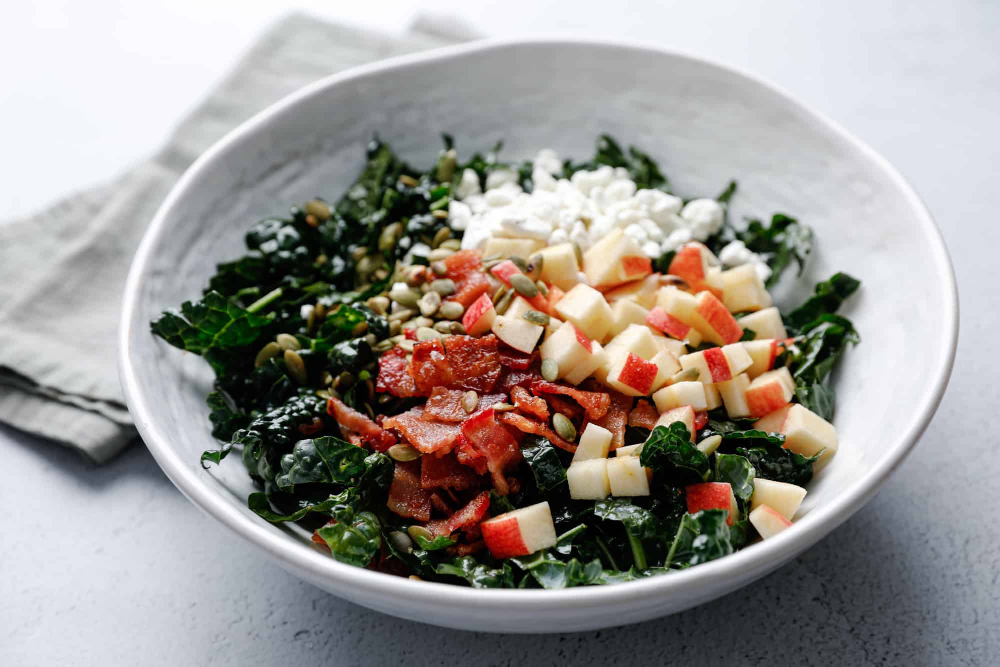 A white bowl filled with kale salad topped with goat cheese, bacon, pepitas, and apple