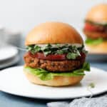 Two veggie burgers on white plates on a blue background with toasted buns, green sprouts, butter lettuce and heirloom tomato.