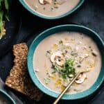 Overhead view of three bowls of cream of mushroom soup with garnish, a spoon and a side of seed crackers