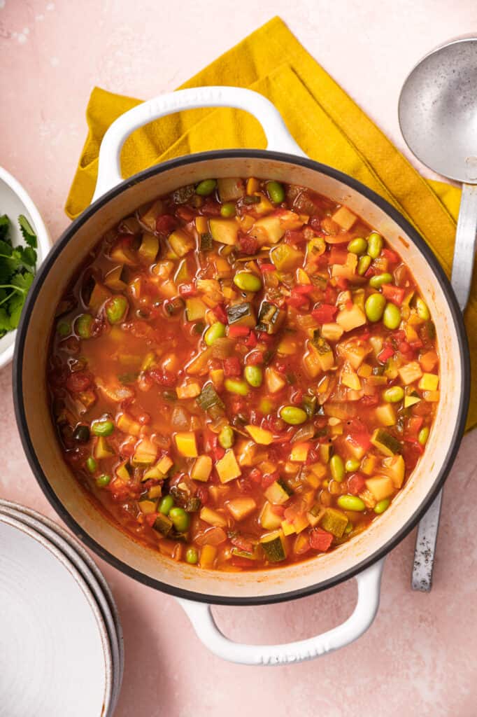 Overhead view of a pot of succotash with lots of veggies on top of a yellow napkin and pink background