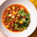 Overhead view of a white bowl of succotash with lots of veggies on top of a yellow napkin and pink background