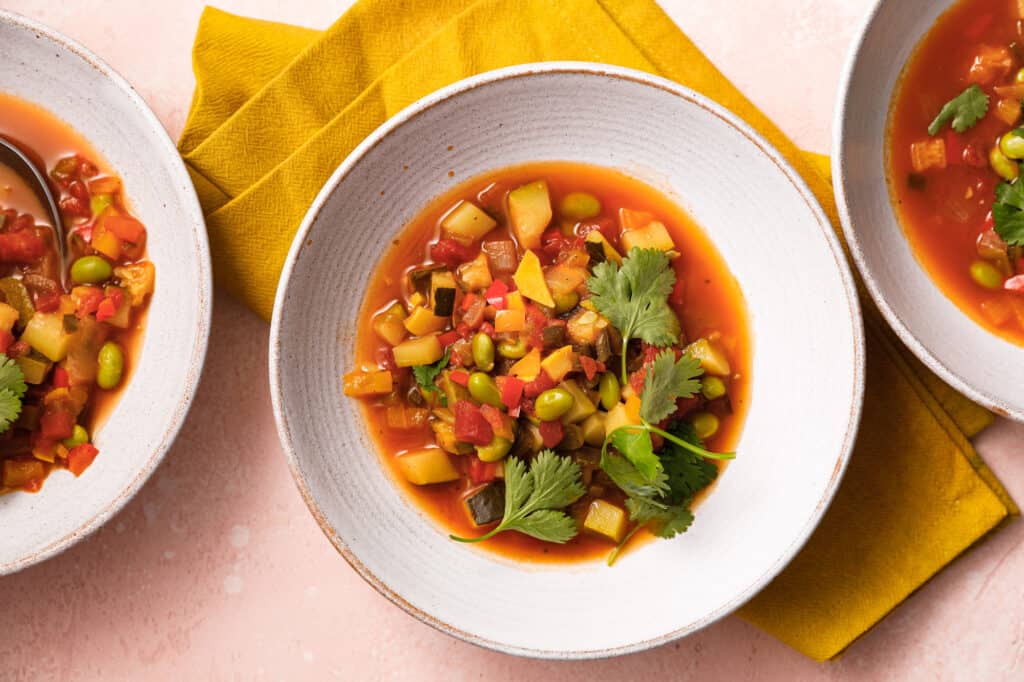 Overhead view of three white bowls of succotash with lots of veggies on top of a yellow napkin and pink background