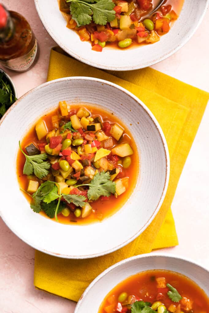 Overhead view of three white bowls of succotash with lots of veggies on top of a yellow napkin and pink background