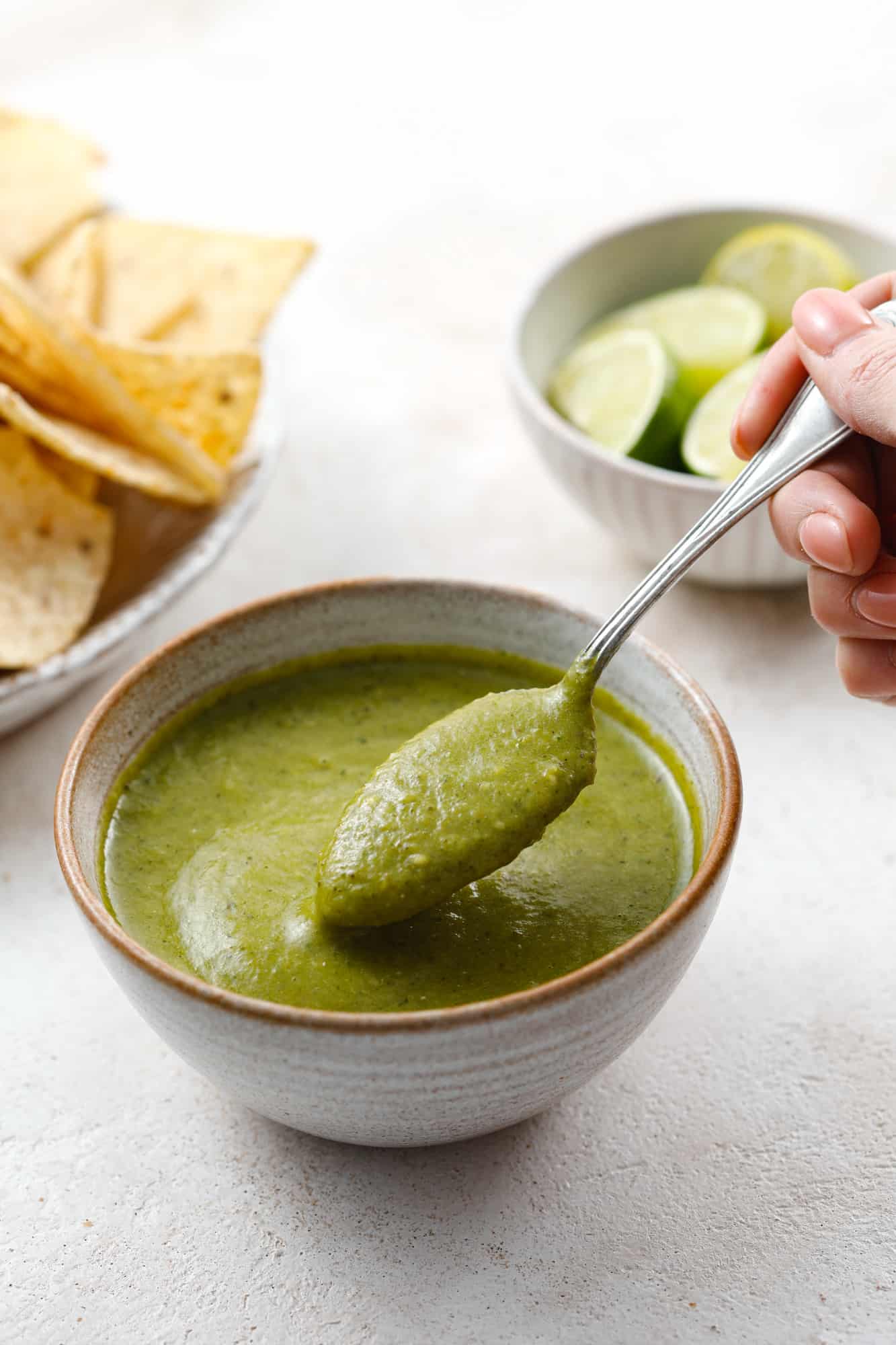 A bowl of bright green jalapeno salsa in a ceramic bowl being scooped with a teaspoon.