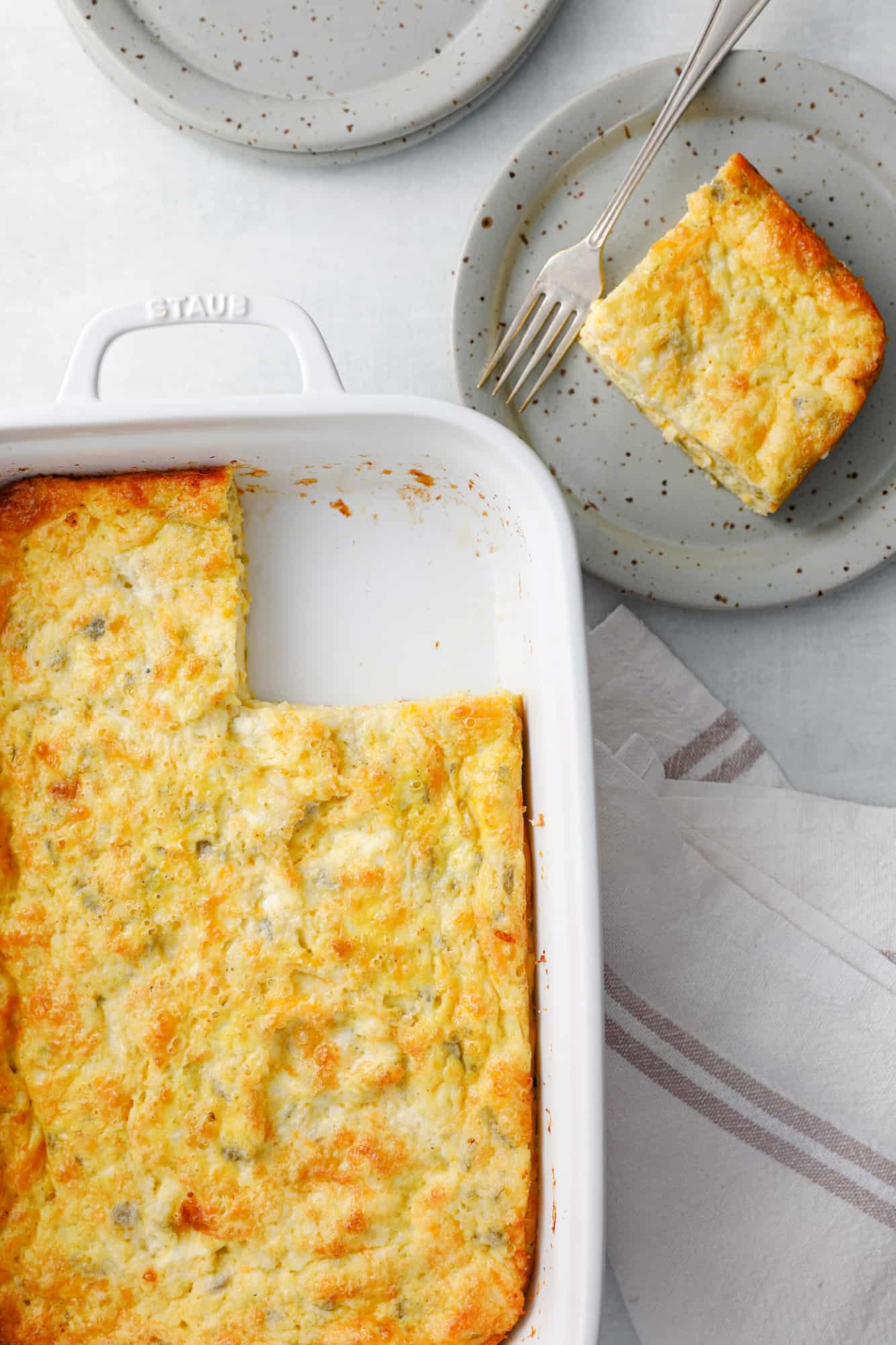Slice of overnight breakfast green chili egg casserole on a grey dish with a fork next to a dish full of more egg casserole