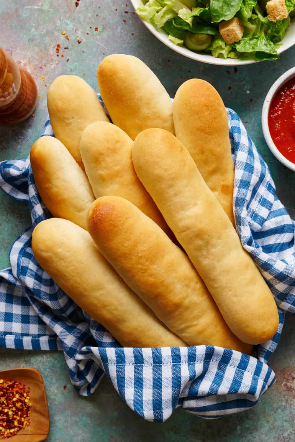 overhead view of breadsticks in a basket with gingham napkin with salad and marinara on the side.