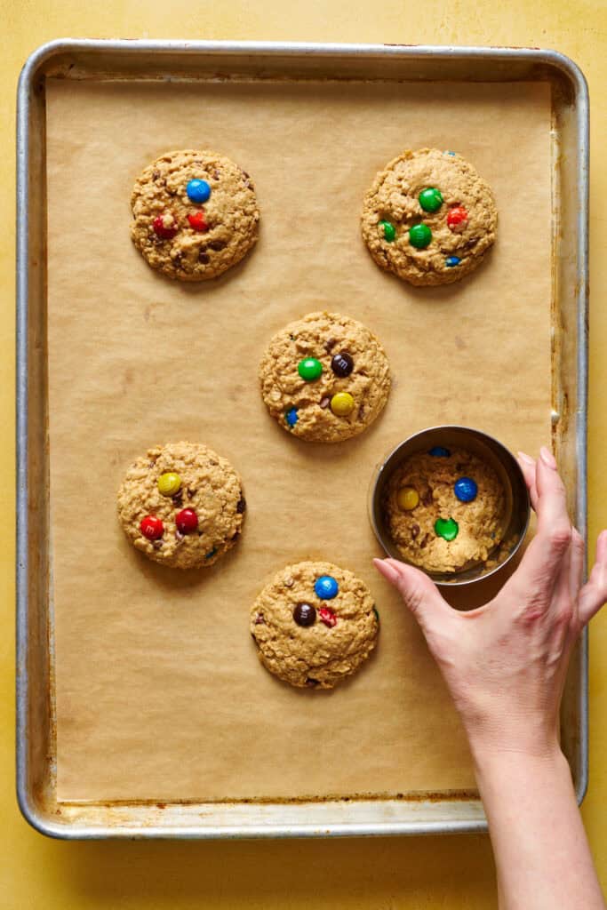 baked monster cookies on a sheet tray and a hand making them round with a 4'' cookie cutter