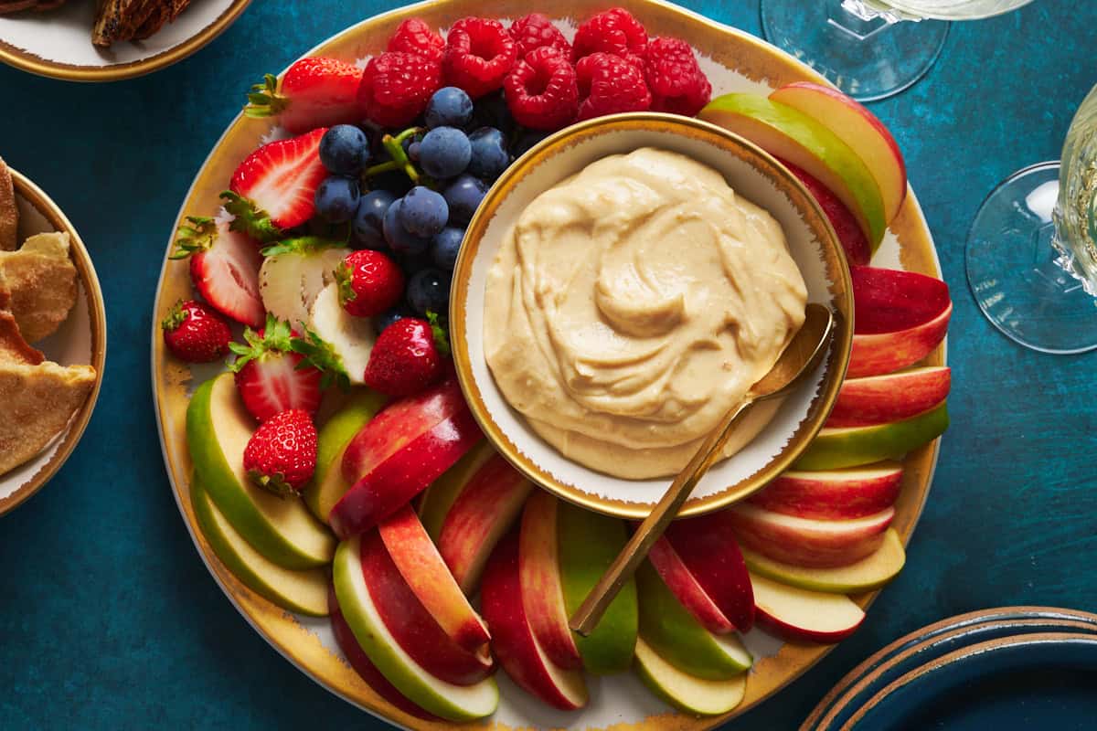 Platter of fruit with cheesecake fruit dip surrounded by plates and drinks as well as sweet crackers for dipping.