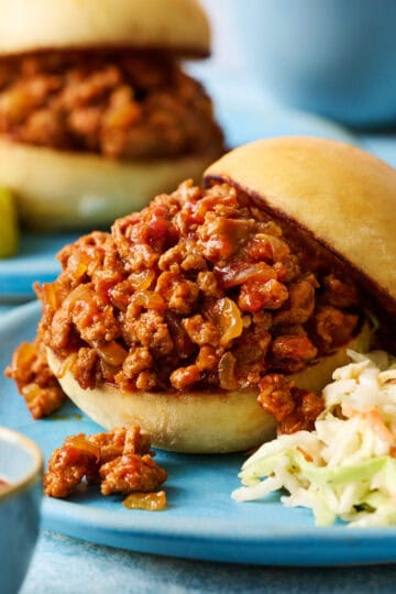 A ground chicken sloppy joes sandwich on a blue plate with coleslaw with another sandwich in the background and cheese in a bowl.