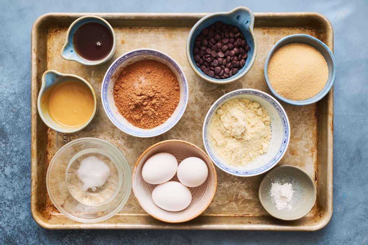 ingredients laid out for making paleo brownies including chocolate chips, almond flour, sugar alternative, baking powder, eggs, coconut oil, cacao, peanut butter and maple syrup