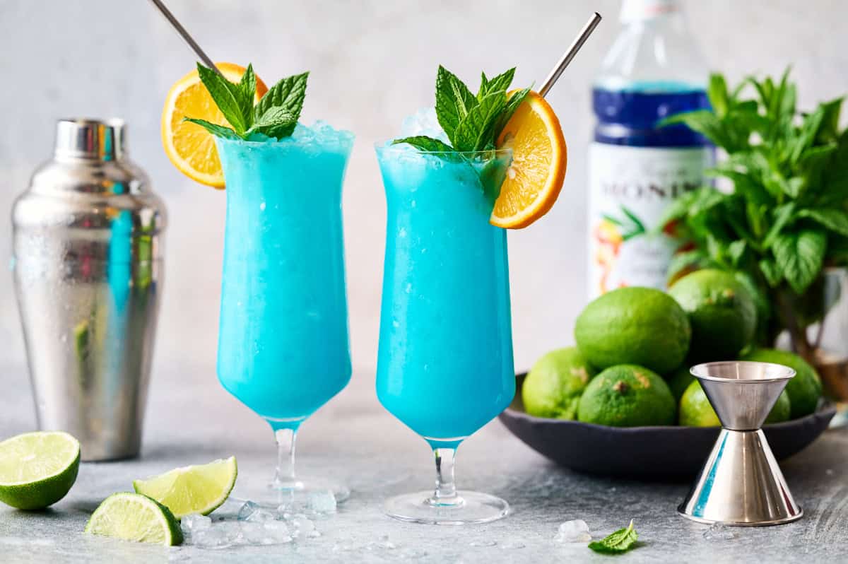 Blue ocean mocktails in hurricane glasses garnished with mint and orange with limes and curacao syrup in the background