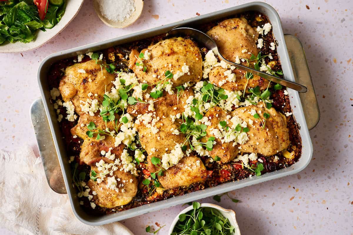 One pan Mediterranean chicken quinoa bake with golden brown chicken thighs, crumbled feta cheese, and fresh herbs in a baking dish, with a serving spoon and side salad visible