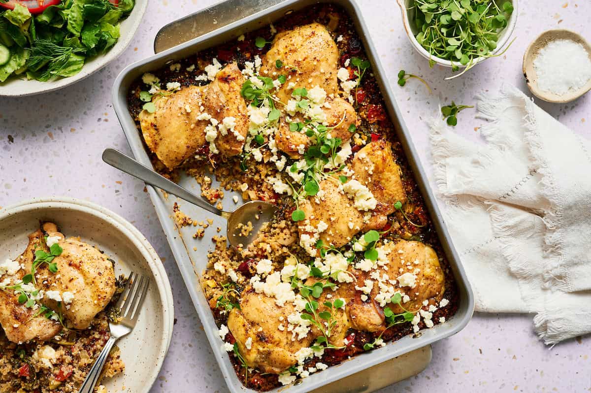 One pan Mediterranean chicken quinoa bake with golden brown chicken thighs, crumbled feta cheese, and fresh herbs in a baking dish, with a serving spoon and side salad visible