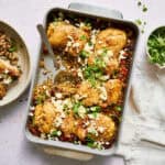 One pan Mediterranean chicken quinoa bake with golden brown chicken thighs, crumbled feta cheese, and fresh herbs in a white ceramic baking dish, with a serving spoon and side salad visible