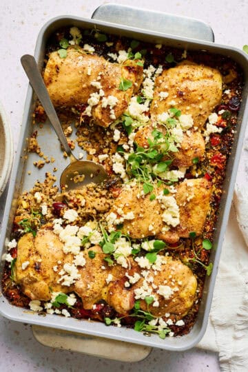 One pan Mediterranean chicken quinoa bake with golden brown chicken thighs, crumbled feta cheese, and fresh herbs in a white ceramic baking dish, with a serving spoon and side salad visible