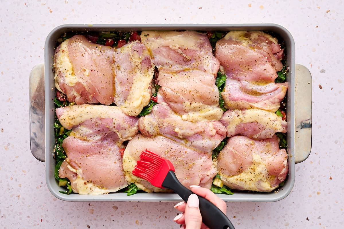 Brushing the seasoned chicken thighs with olive oil in a pan with chicken and vegetables.