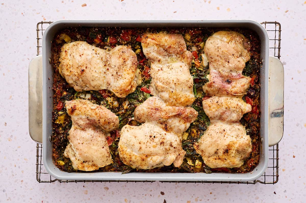 One pan Mediterranean chicken quinoa bake with golden brown chicken thighs, crumbled feta cheese, and fresh herbs in a white ceramic baking dish, with a serving spoon and side salad visible