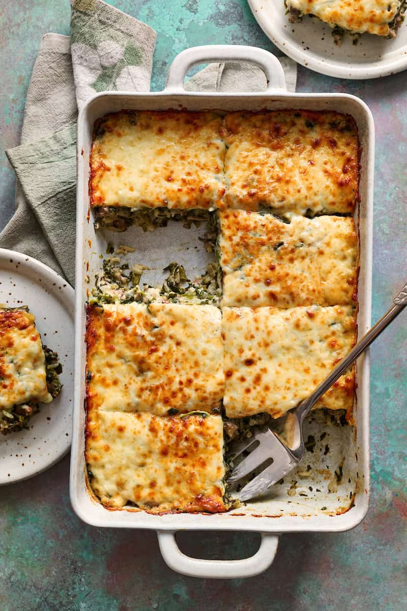 A large dish filled with a spinach, cheese and beef casserole with a piece taken out and put on a plate with a fork