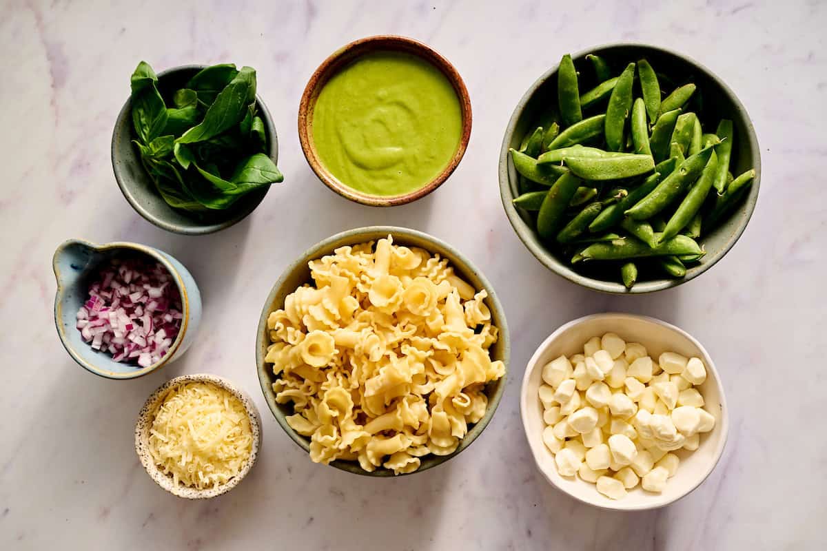 ingredients on a tabletop including snap peas, mozzarella, pasta, parm, red onion and basil