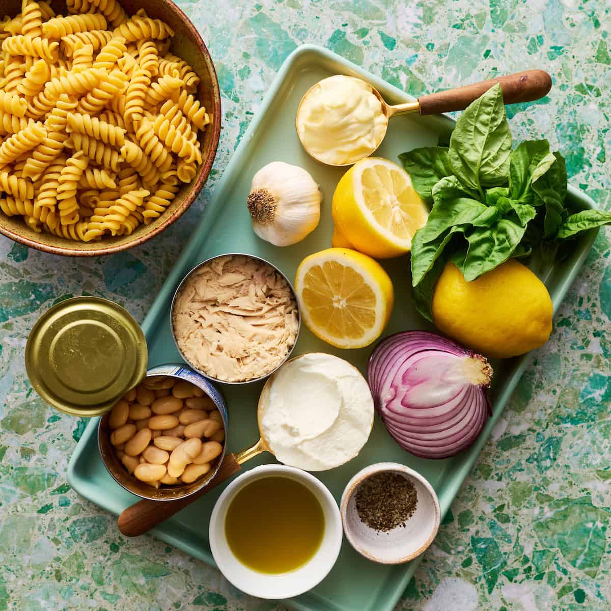 ingredients for making tuna pasta salad including fresh basil, lemon, garlic, onion, olive oil, pasta and tuna