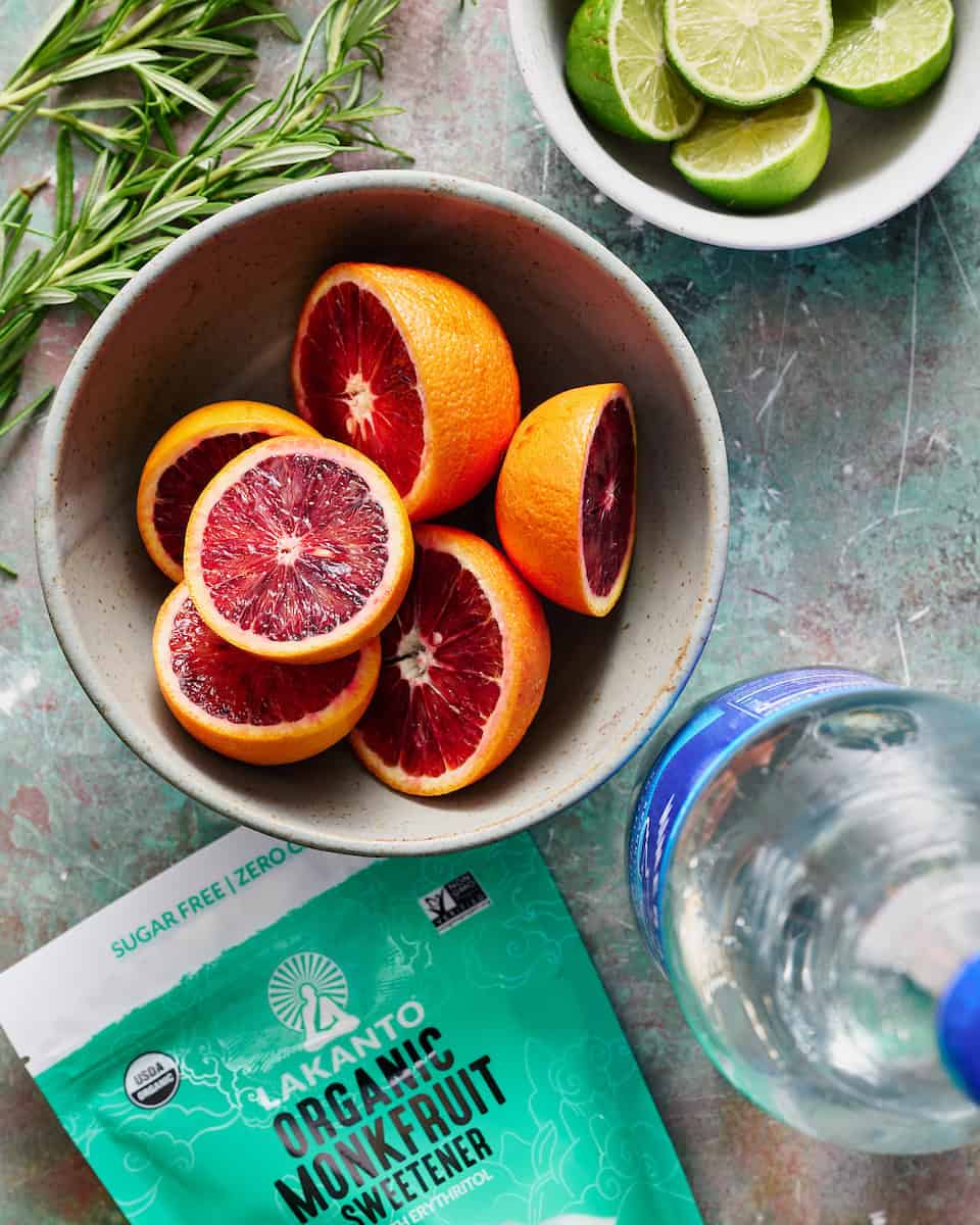 ingredients to make a blood orange mocktail including rosemary, sparkling water, and limes